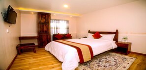 A bedroom with a wooden bed, a TV mounted on the wall, a wooden bench, and a patterned rug. - MeroStay 141 Hotel RG (Kathmandu)
