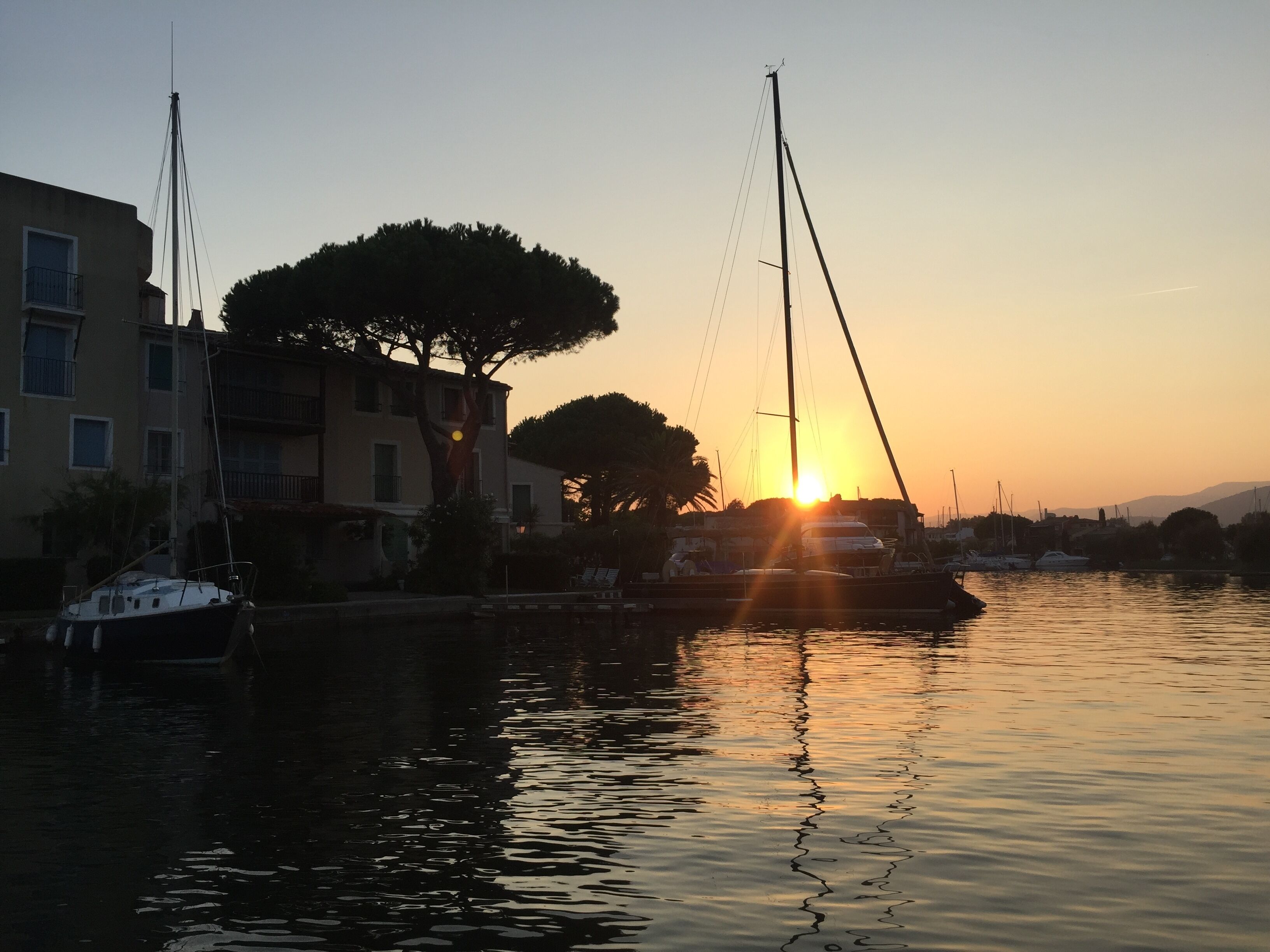 Ein Yachthafen bei Sonnenuntergang mit vor Anker liegenden Booten, Gebäuden und Bäumen.