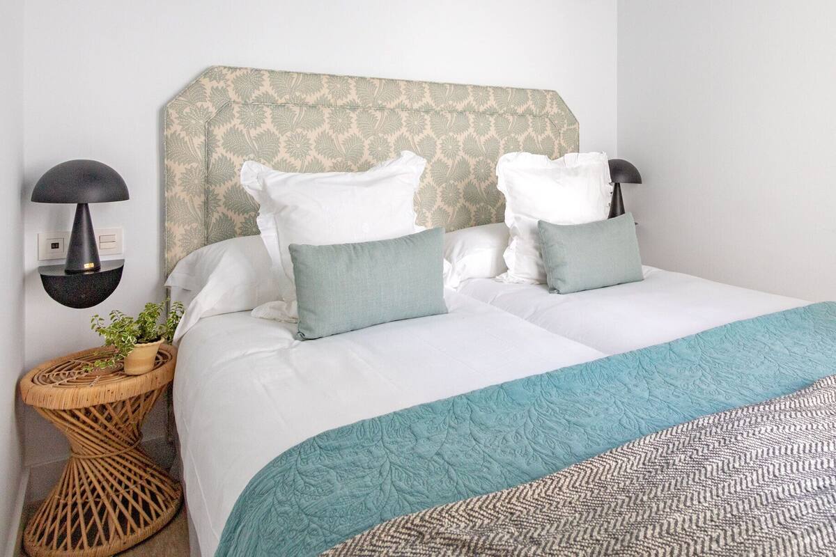A neatly made bed with white linens and teal accents, a wall-mounted lamp, and a woven side table with a plant.