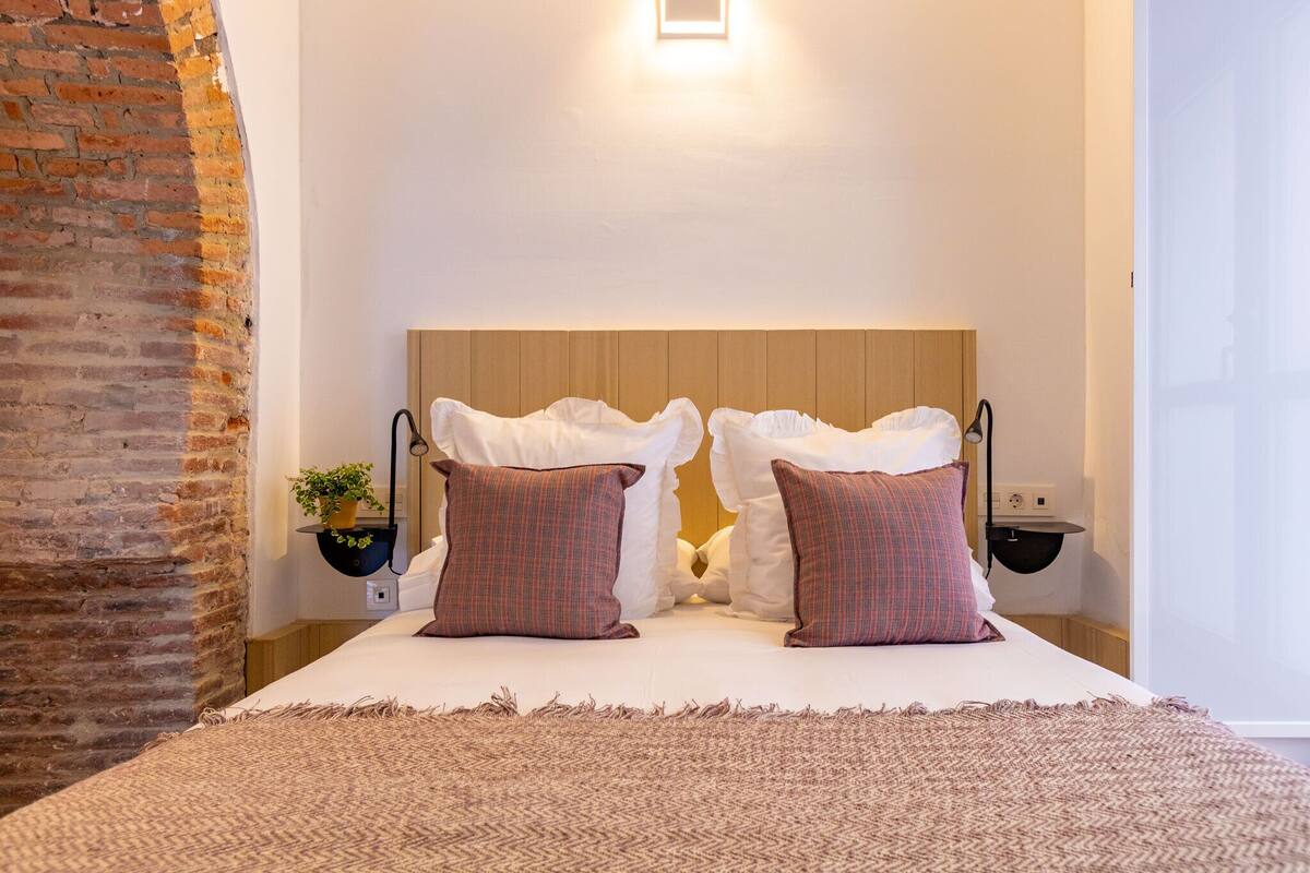 A neatly made bed with white linens and two plaid pillows, a small potted plant on a bedside table, and a brick wall on one side.
