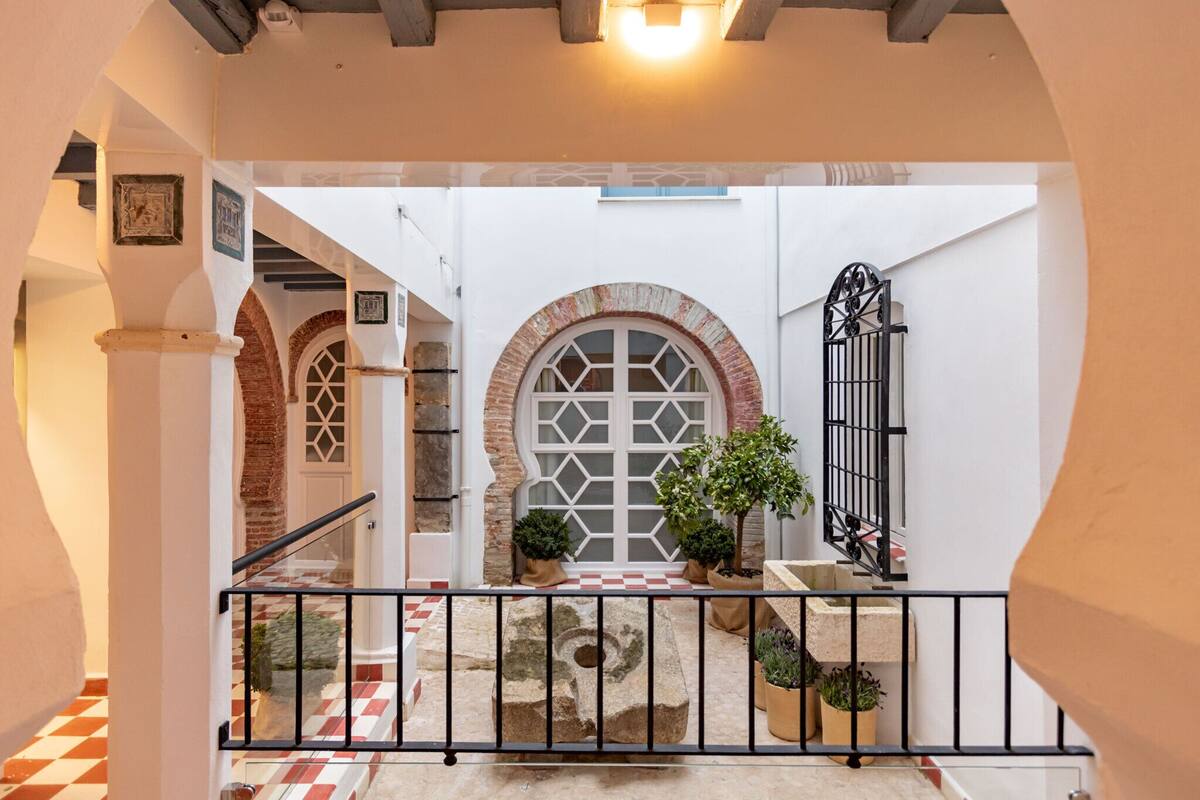 A courtyard with a checkered floor, a stone bench, potted plants, and a metal railing.