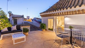 An outdoor patio with a white sofa, a white coffee table, and metal chairs. There is a tiled roof and a staircase leading to an upper level. - Pimienta 9 - Santa Cruz by Valcambre  (Seville)