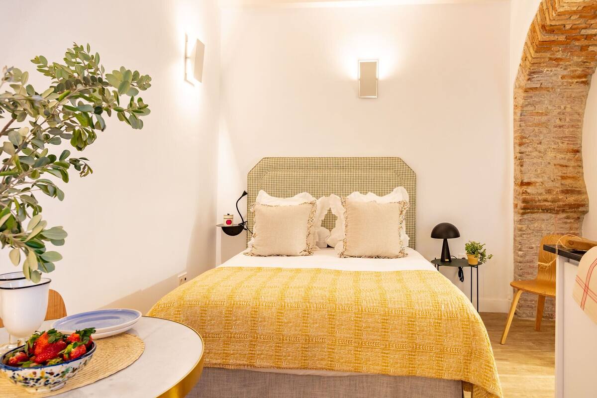 A cozy bedroom with a checkered headboard, a yellow bedspread, a bedside table with a lamp, and a small plant.