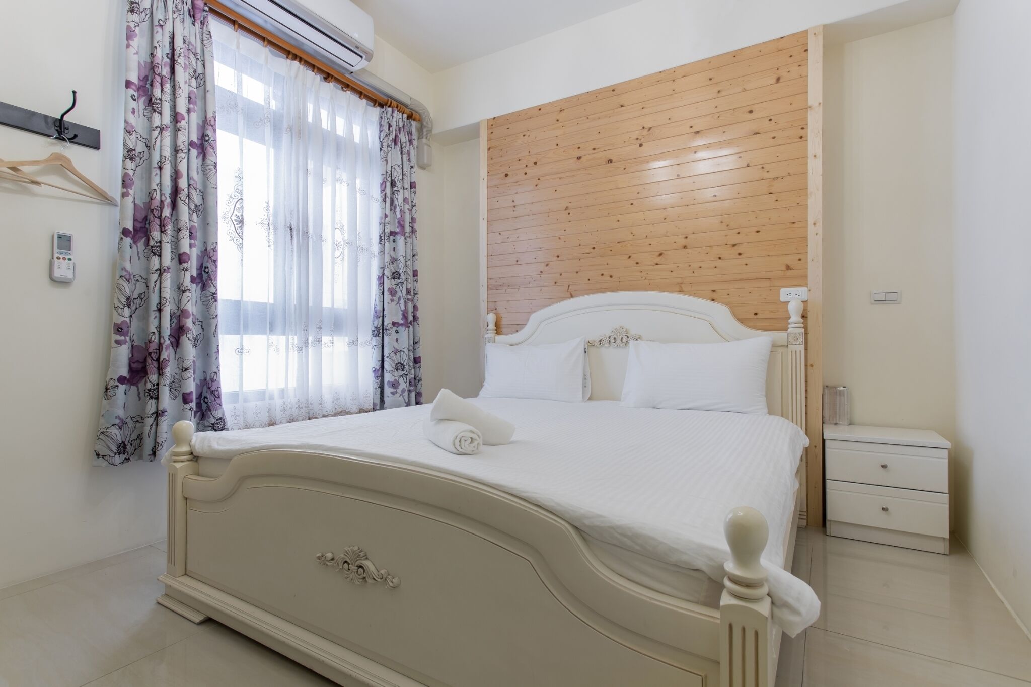 A bedroom with a bed, a nightstand, a wall-mounted air conditioner, and a window with floral curtains.