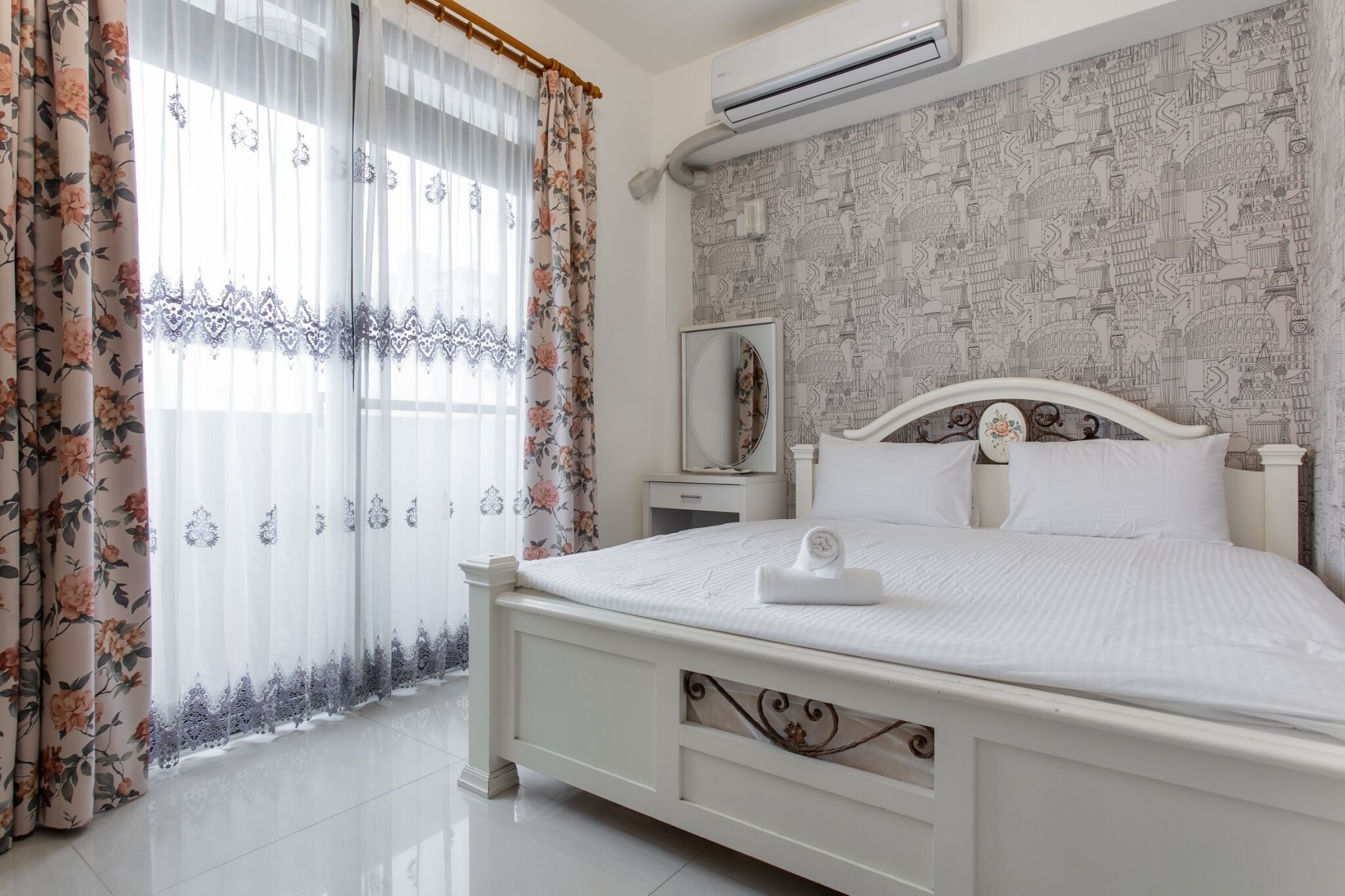 Una cama bien hecha con cabecera blanca, una mesita de noche con espejo y una cortina con estampado floral.