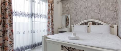 Una cama bien hecha con cabecera blanca, una mesita de noche con espejo y una cortina con estampado floral.
