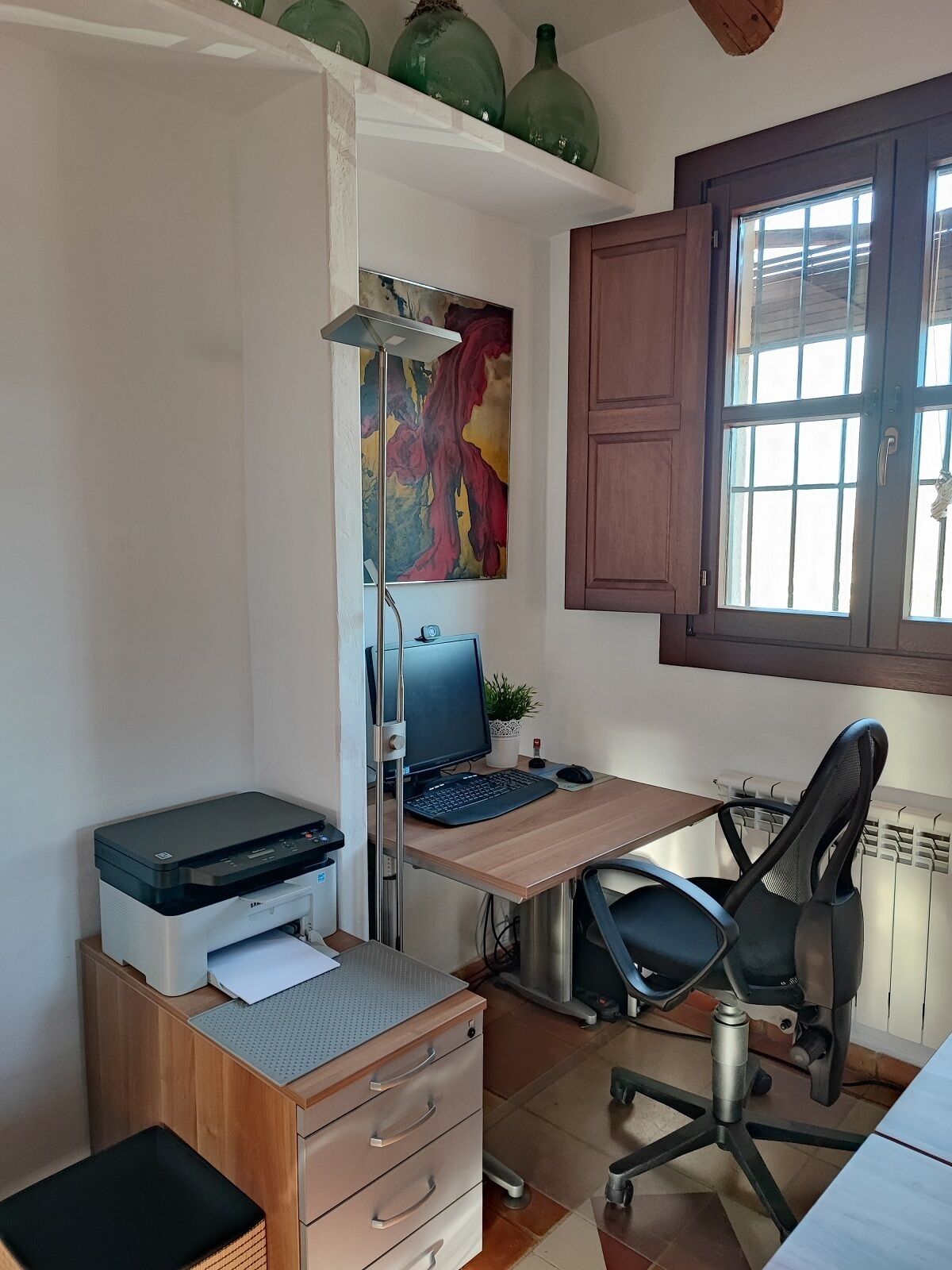 A home office with a desk, computer, printer, and chair. There is a window with a view, a radiator, and a painting on the wall.