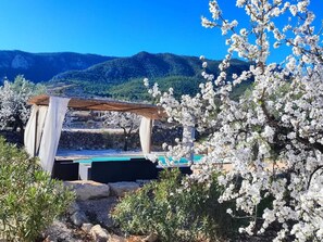 Un espacio de estar rústico al aire libre con una mesa de piedra y cortinas blancas, rodeado de flores blancas en flor y olivos, con montañas al fondo.