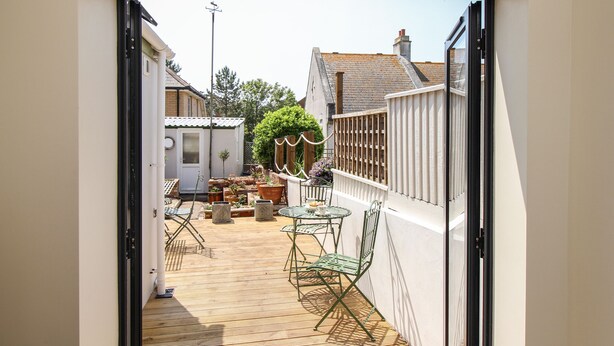 A narrow outdoor patio with a wooden floor, metal chairs, a small table, and potted plants. - Blanche Cottage (Weymouth)