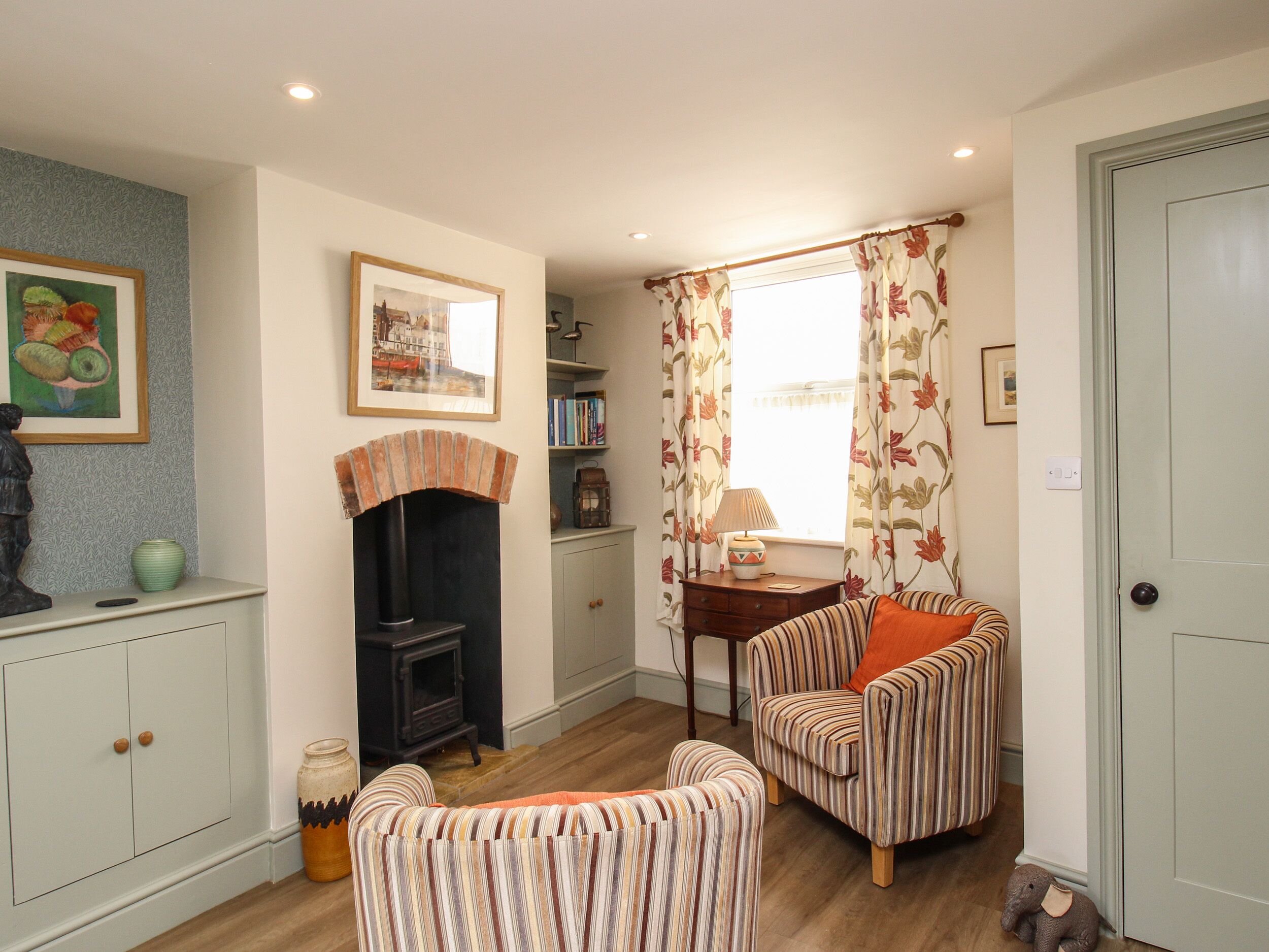 A cozy living room with a fireplace, striped armchairs, a bookshelf, and a window with floral curtains.
