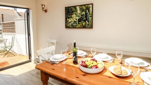 A dining area with a wooden table set for two, a fruit bowl, wine glasses, and a bottle of wine. A painting hangs on the wall, and there is a view of a garden outside. - Blanche Cottage (Weymouth)
