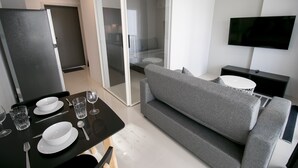 A modern living room with a grey sofa, a dining table set with plates and glasses, and a flat-screen TV mounted on the wall.