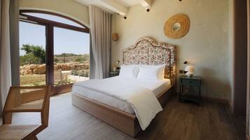 Un dormitorio con una cama grande, una silla de mimbre, una mesita de noche con lámpara y vistas al exterior.