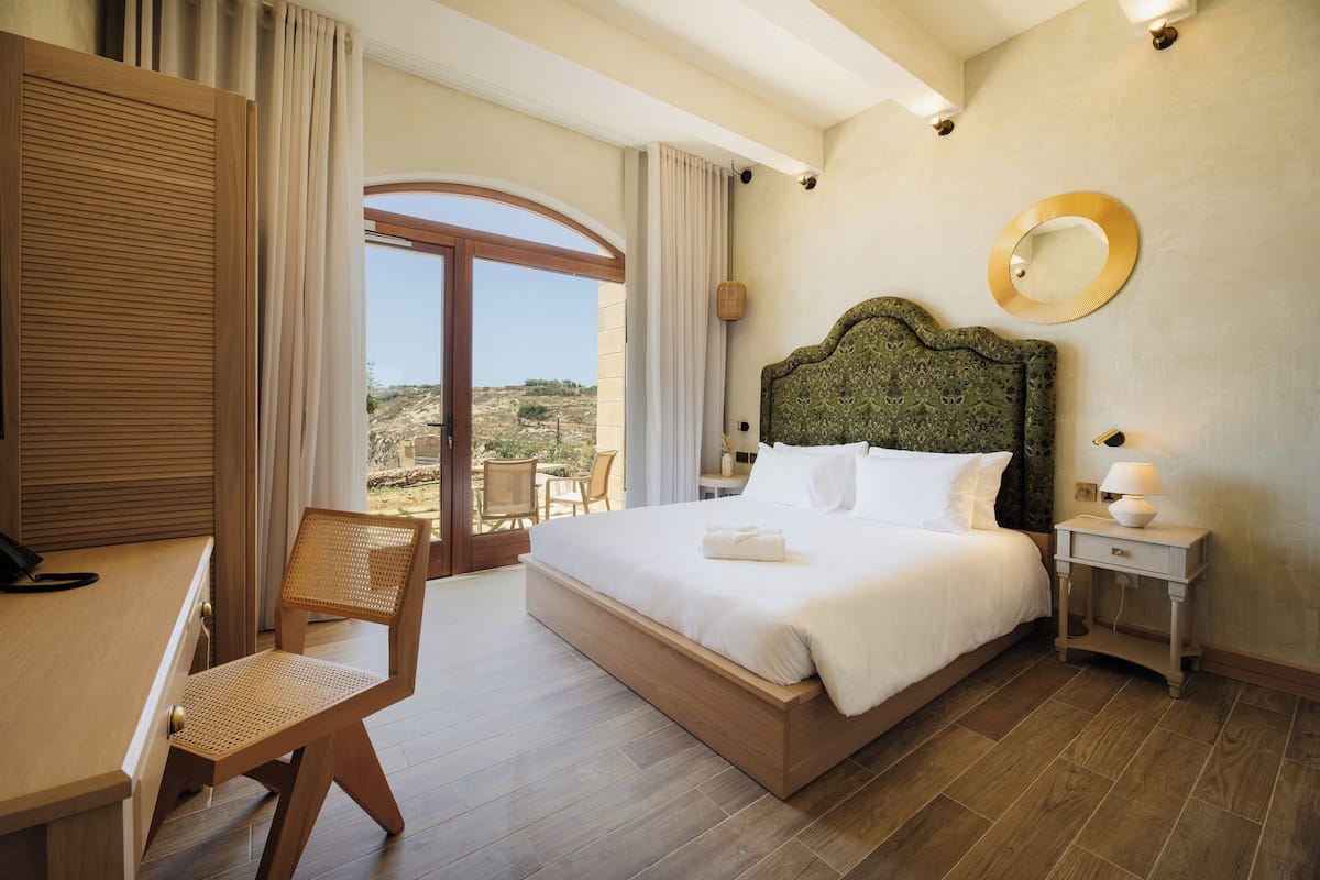 A bedroom with a large bed, a wooden desk, a chair, a nightstand with a lamp, and a view of the outdoors.