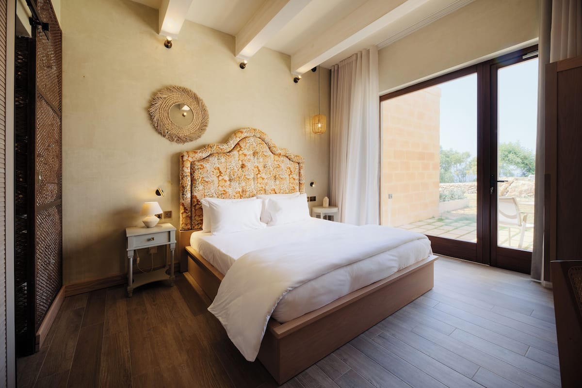 A bedroom with a large bed, a nightstand with a lamp, a decorative wall hanging, and a view through a sliding door to an outdoor area.