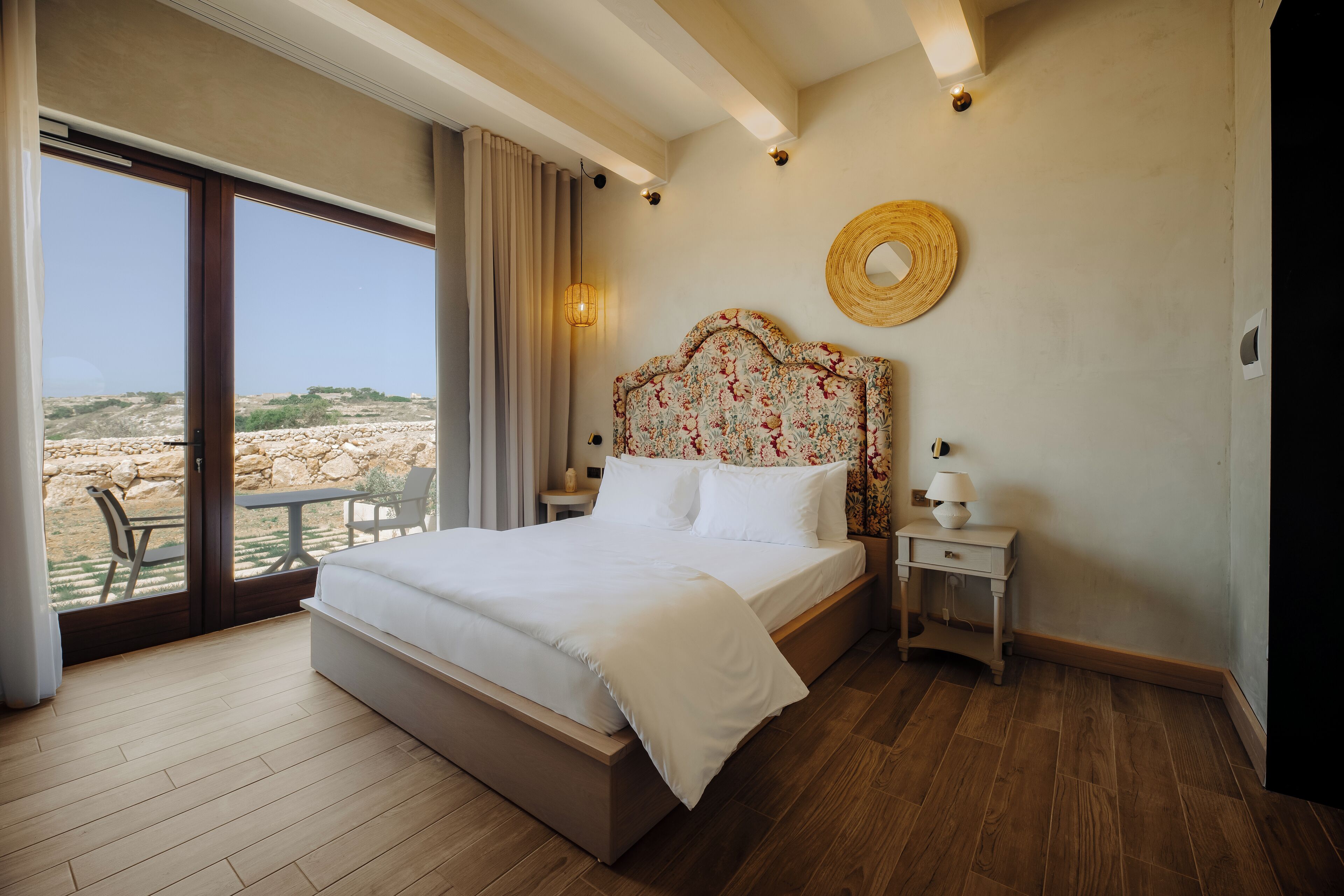A bedroom with a large bed, a nightstand, a lamp, and a view of the outdoors.
