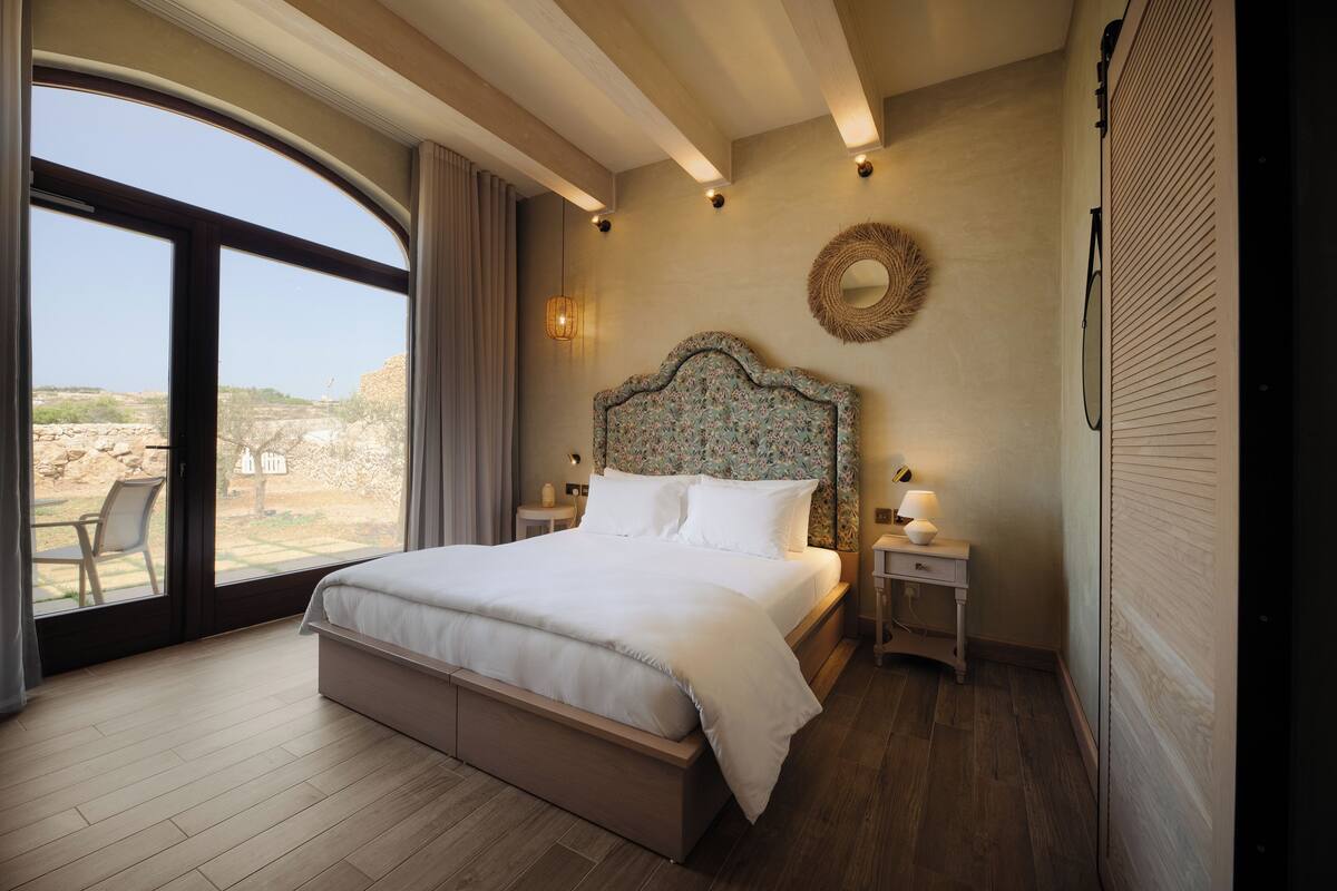 A bedroom with a large bed, a view of the outdoors, a round mirror, and a small bedside table with a lamp.