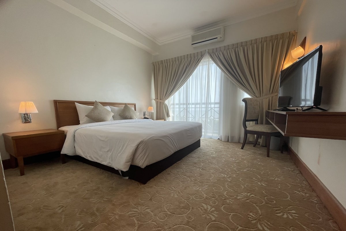 A hotel room with a large bed, a TV, a desk, and a window with curtains.