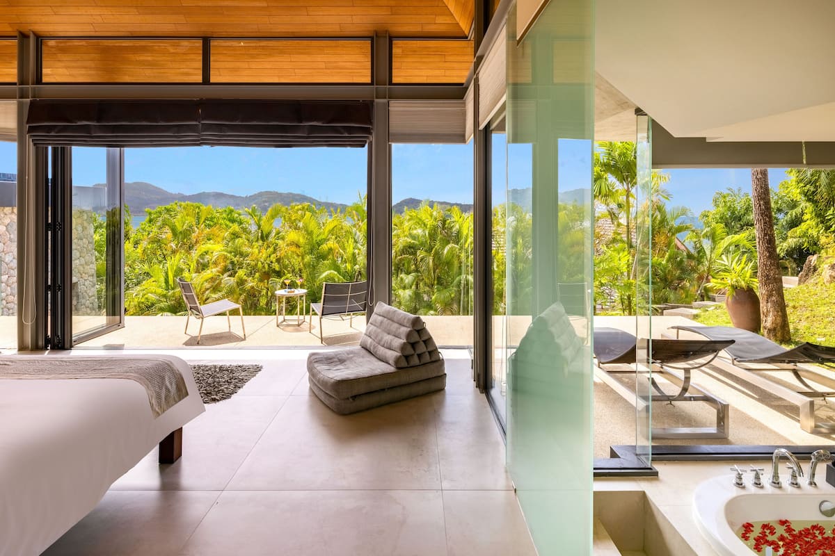 Quarto moderno com uma cama grande, um sofá e vista para um jardim exuberante e montanhas.