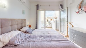 Een slaapkamer met een bed, nachtkastje, lamp, kast en balkon met uitzicht op gebouwen.
