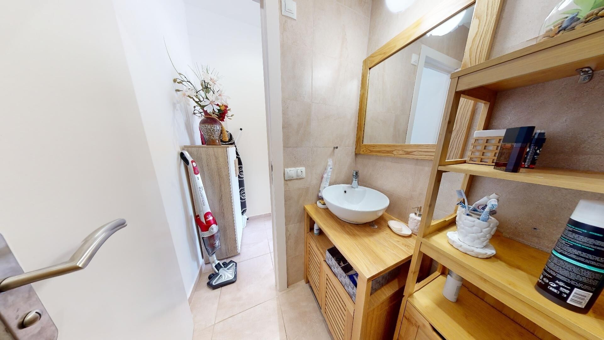 A modern bathroom with a wooden vanity, a large mirror, and a sink. There is a shelf with toiletries and a vacuum cleaner in the corner.