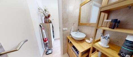 A modern bathroom with a wooden vanity, a large mirror, and a sink. There is a shelf with toiletries and a vacuum cleaner in the corner.