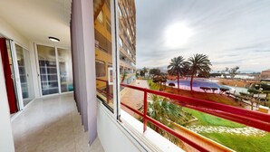 A balcony with a red railing overlooking a pool area with palm trees and a building in the background. - Bel Appartement Avec Piscine , le 600 (ROSES)