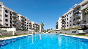A swimming pool surrounded by modern apartment buildings with balconies, a clear blue sky, and a palm tree in the background. - Global Vacacional: Apartment with large terrace in Canet (Canet d´en berenguer)