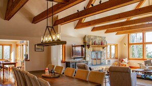 A spacious living room with a large wooden dining table, comfortable seating, a stone fireplace, and large windows offering a view of the outdoors. - Mountainside 5-BR Ski-In/Out Okemo Retreat (Ludlow)