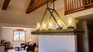 A cozy living room with a wooden ceiling, a hanging light fixture with multiple bulbs, a sofa, a small table, and a window with a view. - Mountainside 5-BR Ski-In/Out Okemo Retreat (Ludlow)