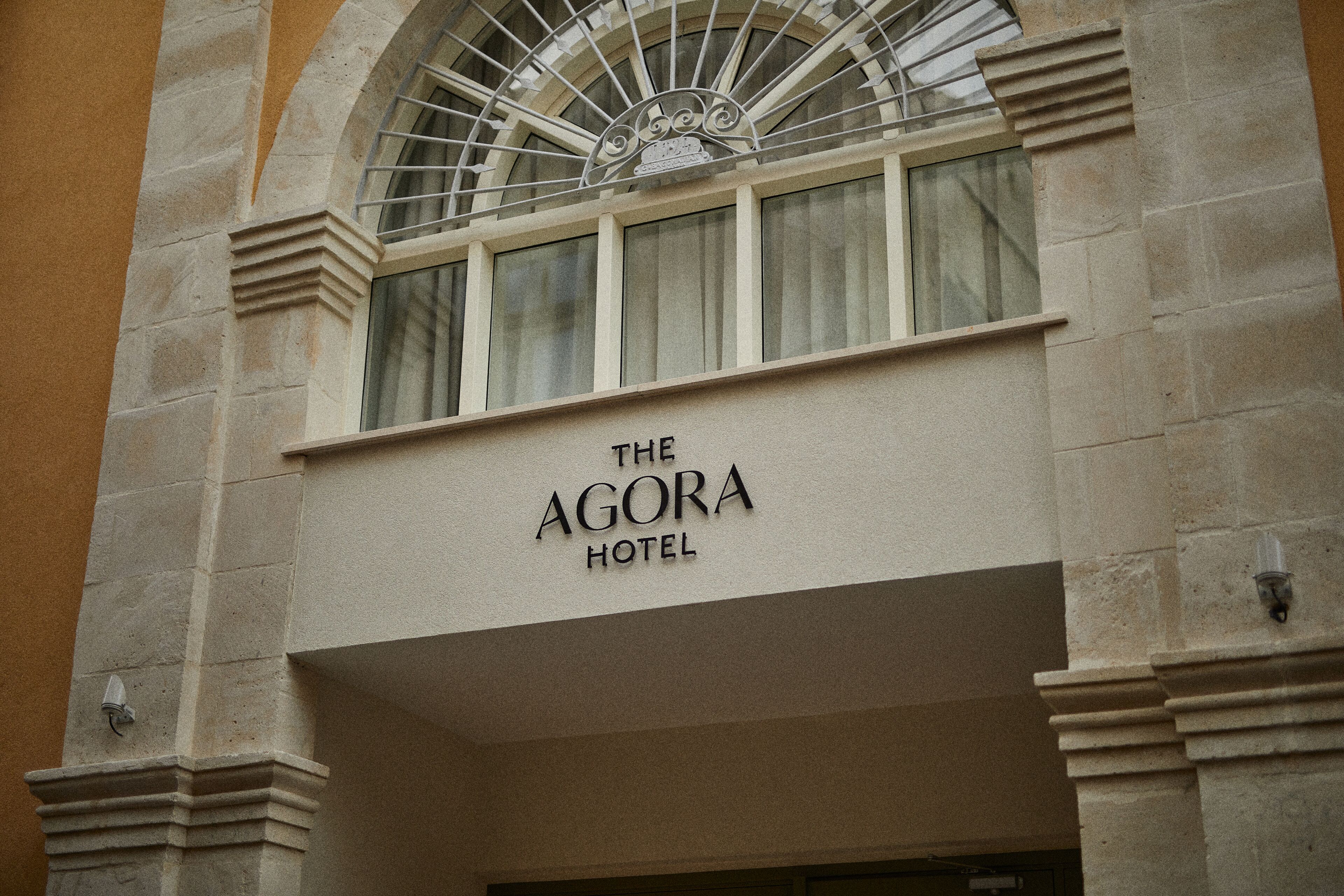 Die Fassade des Agora Hotels mit einer auffälligen Beschilderung, Steinarchitektur und einem dekorativen Fenstergitter.