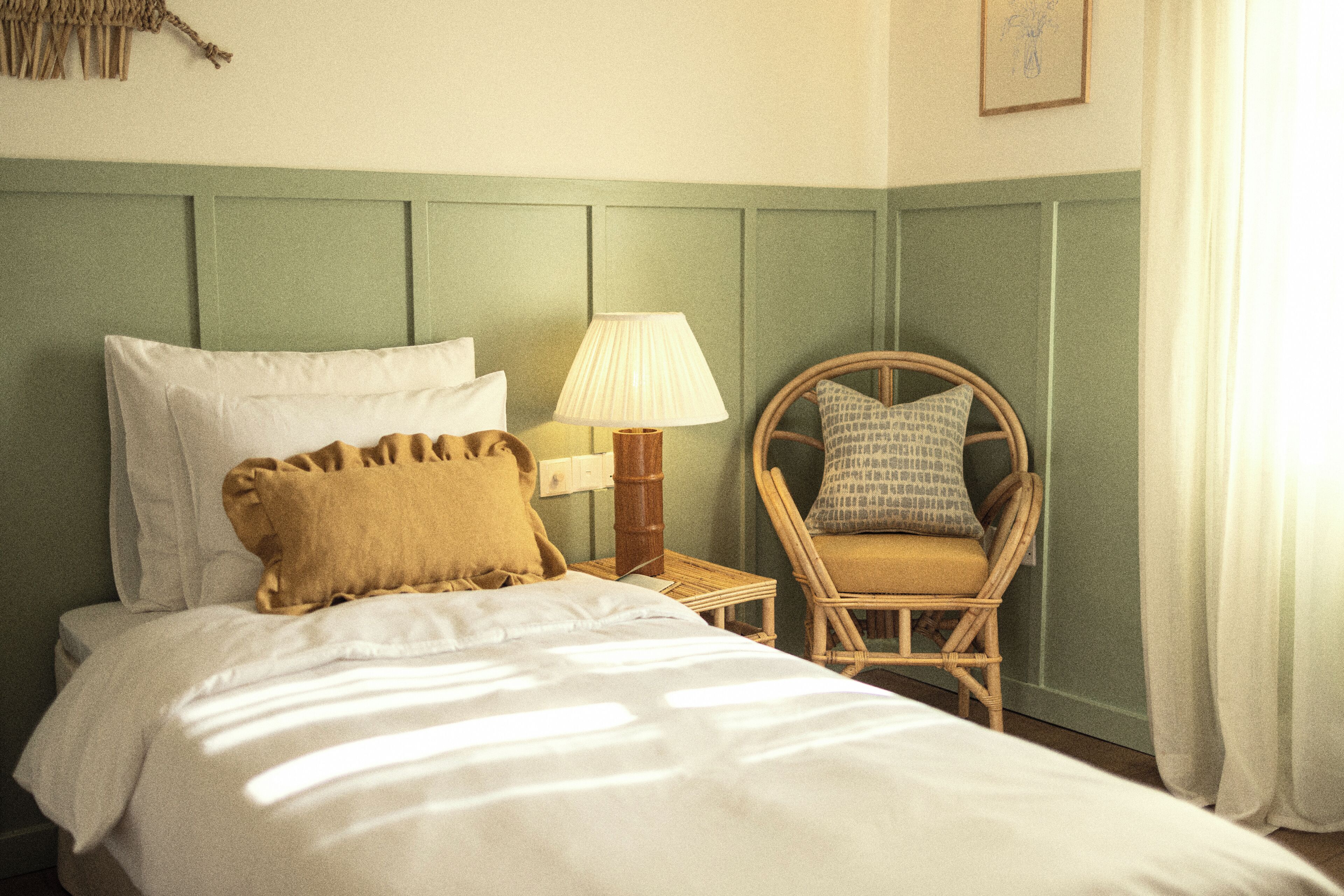 Un lit bien fait, avec du linge de lit blanc et un oreiller marron, une lampe de chevet et une chaise en osier avec un coussin.