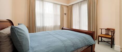 A neatly made bed with a blue bedspread and pillows, a wooden nightstand, a wooden chair, and a window with sheer curtains.