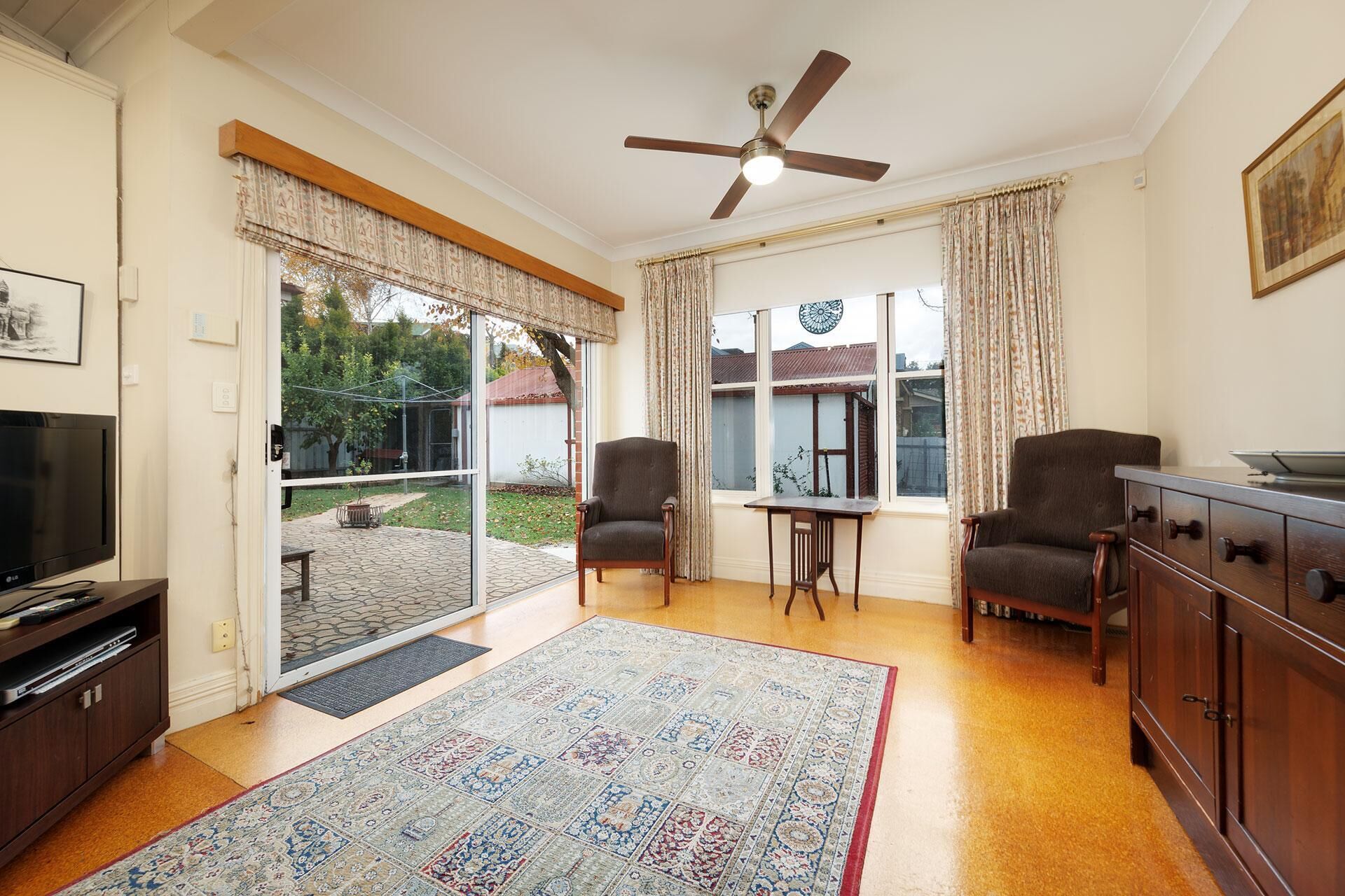 Una sala de estar con televisor, dos sillones, una mesa de centro, un ventilador de techo y un ventanal con cortinas.