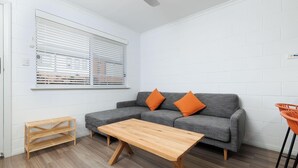 A modern living room with a grey sofa, two orange cushions, a wooden coffee table, a small wooden side table, and a window with blinds. - Charlie's on Stanley - Cosy one bedroom apartment in the centre of Albury (Albury)