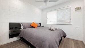 A bedroom with a bed, a bedside table, a ceiling fan, a window with blinds, and a white brick wall. - Charlie's on Stanley - Cosy one bedroom apartment in the centre of Albury (Albury)