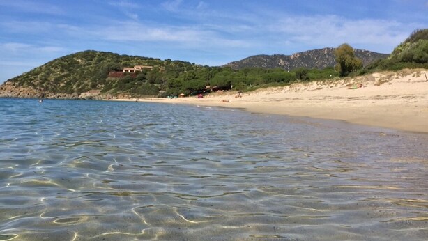 Ein Strand mit klarem, flachem Wasser, sandigem Ufer und einer Hügelkulisse mit Vegetation und Gebäuden im Hintergrund.