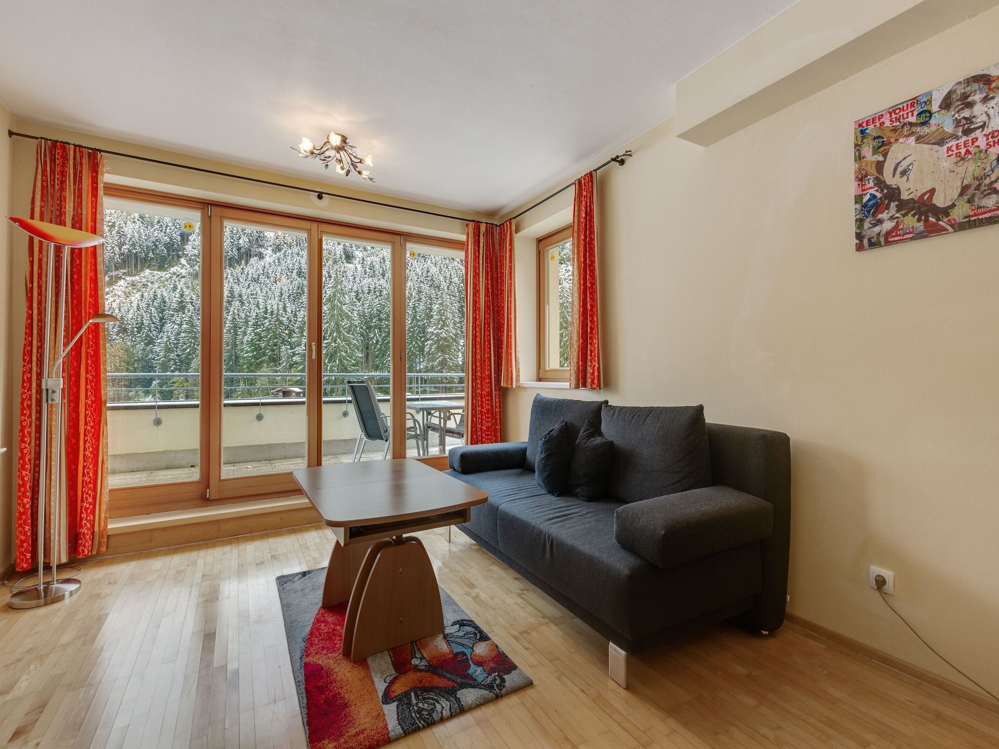 En moderne stue med sofa, sofabord og spisebord. Store vinduer med gardiner, en lysekrone og et billede på væggen.