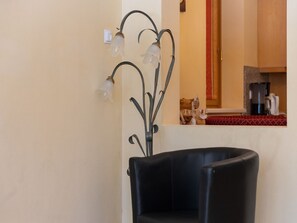 A room with a black leather chair, a wall-mounted light fixture, and a kitchen area visible through an open doorway.