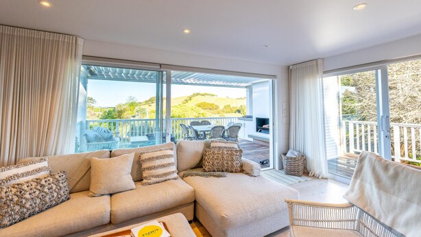 A modern living room with a large sectional sofa, a wicker chair, and a view of a deck and outdoor area. - Oriwa Villa, Oneroa Village - near Mudbrick and Cable Bay - Welcome to Oriwa Villa, a luxurious 4 bedroom, 3 bathroom family home. This exquisite holiday home is designed to provide the perfect getaway, offering a blend of elegance, modern amenities, (Waiheke Island)