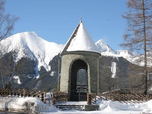 Uma torre de pedra com telhado cônico, entrada em arco e portão de metal, rodeada por neve e montanhas.