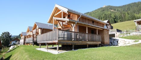 A wooden chalet with a balcony, set in a grassy area with a mountain backdrop.
