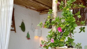 Una zona salotto accogliente con una panca bianca e una poltrona a dondolo in legno, una pianta in vaso con fiori rosa e un soffitto in legno rustico.