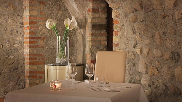Une table à manger dressée, ornée d’un vase de fleurs blanches, de bougies et de verres à vin, dans une pièce aux murs de pierre.