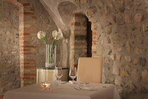 A dining table set with a vase of white flowers, candles, and wine glasses in a stone-walled room. - An exclusive retreat with breathtaking lake views in the hills surrounding Lake Garda (SOIANO DEL LAGO)