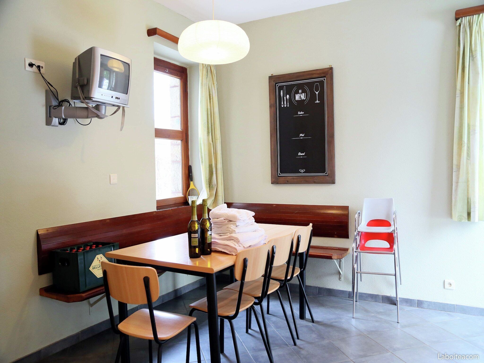 A dining area with a wooden table, chairs, a television mounted on the wall, a chalkboard menu, and a window with curtains.