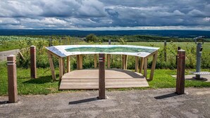 Une plateforme d’observation en bois, agrémentée d’une carte, entourée d’une clôture et dotée d’un télescope, offrant une vue panoramique sur un paysage composé de champs et de collines lointaines.