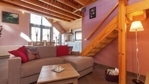 A cozy living room with a wooden ceiling, a sofa with red cushions, a small table, and a staircase. - La Roche Cozy Forest Escape (Marcouray)
