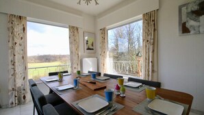 Ein Esszimmer mit einem langen Tisch, gedeckt für sechs Personen, großen Fenstern mit Vorhängen, einer Kristallleuchter und einem Gemälde an der Wand.