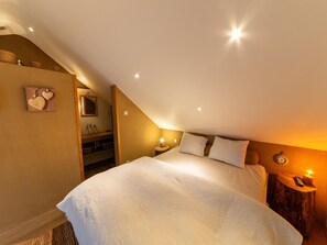 A bedroom with a large bed, a wooden headboard, a nightstand, a bedside table with a lamp, and a wall-mounted clock. - Bertrix charming 2-bedroom holiday home (Bertrix)
